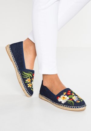 Fødder iført hvide bukser og marineblå espadrillos med farverige broderede blomster og såler af jute på en ens hvid baggrund.