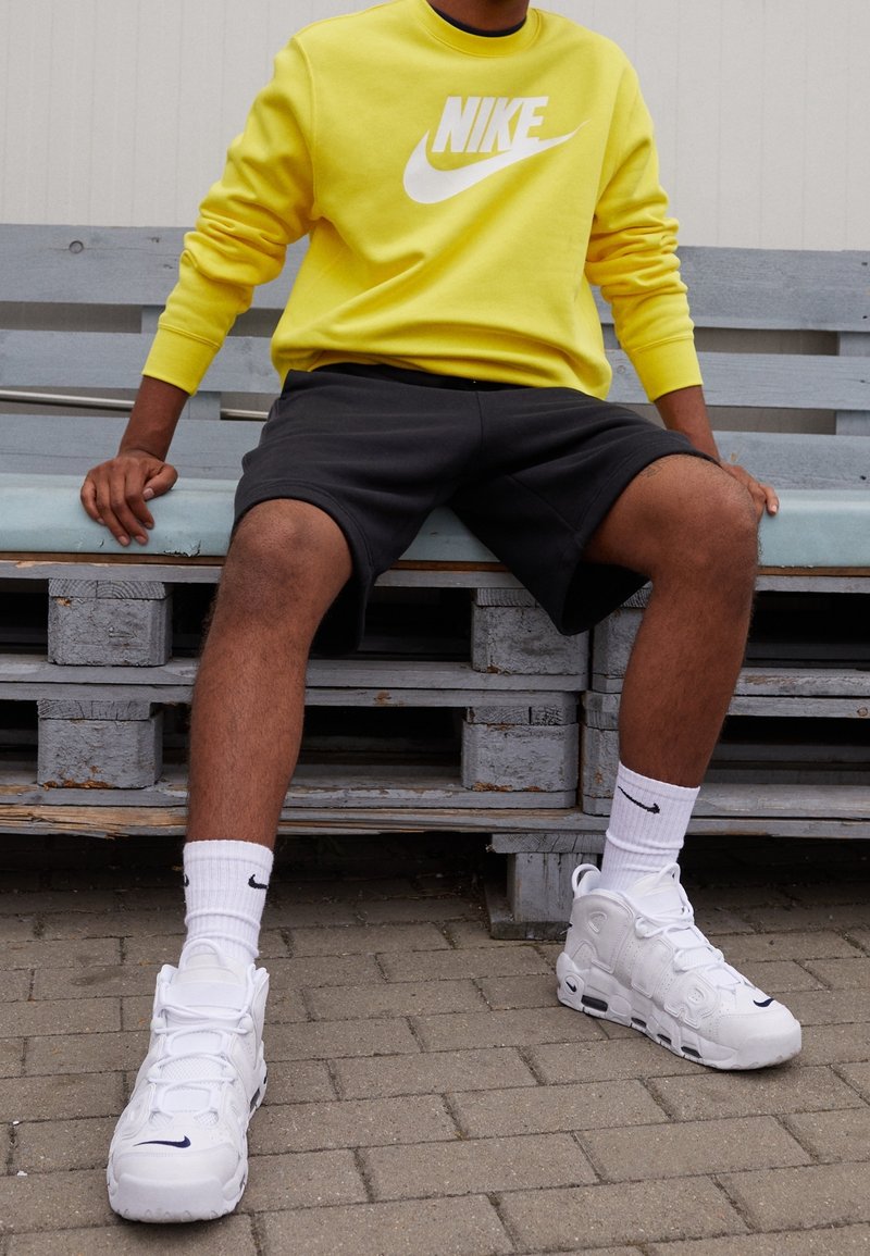 Nike gul sweatshirt med vit logotyp, svarta shorts, vita strumpor med svart logotyp och vita sneakers, sittande på en blå träbänk.