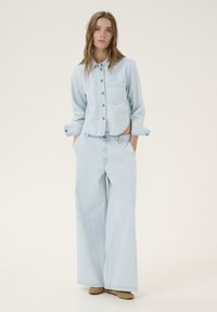 Light blue denim shirt with a chest pocket and button closure, paired with wide-leg matching denim pants and brown shoes, on a neutral background.