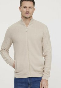 Pull zip beige avec un tissage texturé, coupe ajustée, manches longues et poches latérales. Tissu lisse avec un détail de logo de marque subtil.