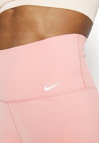 Leggings de color rosa claro de talle alto, confeccionados con tela elástica, con un logo blanco de Nike en la parte frontal y costuras planas para mayor comodidad.