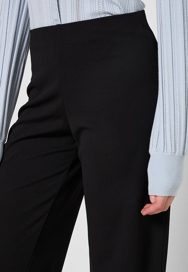 Pantaloni neri a vita alta con una texture liscia, abbinati a una maglietta a maniche lunghe rigata di un azzurro chiaro, con strisce verticali e maniche a polsino.