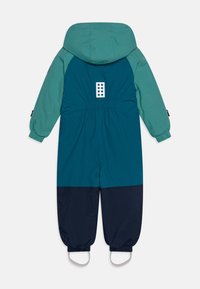 Barevný dětský snowsuit, který má tyrkysový vršek, tmavě modrou spodní část, elastické manžety, kapuci a detail loga na zadní straně.