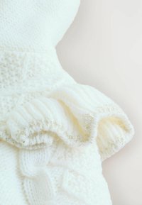 Weißes gestricktes Kleidungsstück mit einem strukturierten Zopfmuster und Rüschenkanten. Die Rüschen sind mit metallischen Fadendetails akzentuiert.