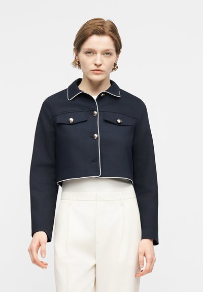 Femme portant une veste courte bleu marine avec des boutons argentés et des bords blancs soulignés, associée à un pantalon blanc taille haute et des boucles d'oreilles créoles dorées.
