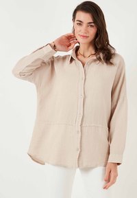 Chemise beige clair à boutons avec un tissu texturé, des épaules tombantes et un ourlet arrondi. Dotée d'un col classique et de détails de couture subtils.