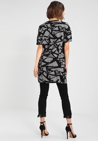 Černá a šedá tunika s krátkými rukávy se vzorem, spárovaná s černými skinny kalhotami s třásněmi na lemech a černými sandály na vysokém podpatku.