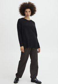 Schwarzer Strickpullover mit geripptem Saum, mit weitem Ärmel. Kombiniert mit locker sitzenden, dunkelbraunen Cargo-Hosen und glänzenden schwarzen Schuhen.