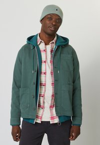 Grüne Jacke mit Knopfverschluss, zwei Vordertaschen und einer Kapuze, über einem karierten Hemd in Rot- und Weißtönen getragen. Graue Strickmütze.