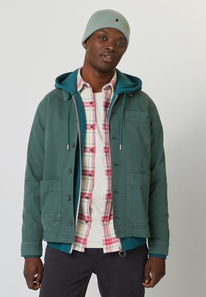 Groene jas met knoopsluiting, twee steekzakken, en een capuchon, gelaagd over een geruite shirt in rood- en wittinten. Grijze brei-beanie.