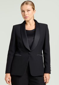 Schwarzer Blazer mit Satin-Schalkragen, Ein-Knopf-Verschluss, zwei Fronttaschen und taillierter Passform; getragen über einem schwarzen Oberteil.