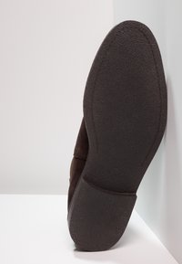 Zapato de ante marrón con suela de goma negra texturizada, que muestra una puntera redondeada y mínimos detalles de diseño. Colocado en posición vertical sobre un fondo claro.