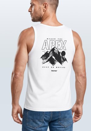 Man wearing white tank top with black mountain graphic and text "Reach the Apex, Keep on Moving" on the back, paired with blue jeans.