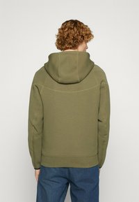 Felpa con cappuccio verde oliva con un grande cappuccio, texture liscia, dettagli di cuciture visibili e vestibilità rilassata, abbinata a jeans di denim blu.