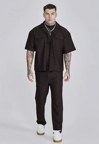 Schwarzes Kurzarmhemd mit vertikalen Streifen und zwei Brusttaschen, kombiniert mit passenden schwarzen Hosen und weißen Sneakern.