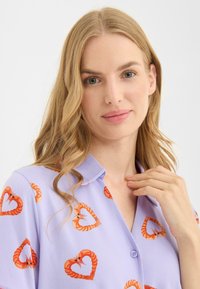 Lilla bluse med mønster af rejer og hjerter, knaplukning foran og korte ærmer. Blødt stof med krave.