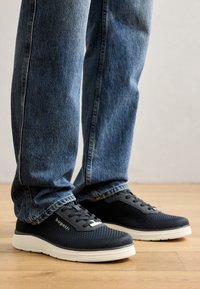 Fekete hálós sportcipők fehér talppal, textúrált felsőrész, fűzős záródás és diszkrét márkajelzéssel az oldalon. Kék farmernadrággal párosítva.