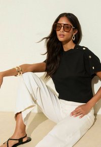 Schwarzes Strickoberteil mit kurzen Ärmeln und Knopfdetails, kombiniert mit weißen weitem Hosen und schwarzen Sandalen. Accessoires umfassen goldene Armreifen und Sonnenbrillen.