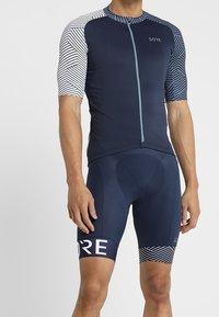 Marinblå cykeldräkt med korta ärmar, med ett geometriskt mönster på axlarna, en framknäppning med dragkedja och förstärkt vaddering i shortsen.