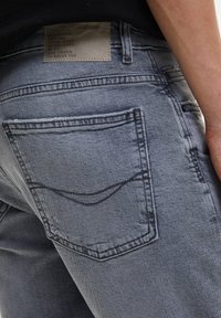 Vaqueros de denim gris con costuras en contraste, que presentan un diseño en el bolsillo trasero y una etiqueta en la cinturilla. La tela parece ligeramente desgastada.