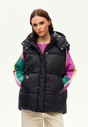 Žena v čiernej prešívanej vesti bez rukávov s kapucňou cez farebný pruhovaný sveter a svetlé nohavice, ruky v vreckách bundy, neutrálny pozadie.