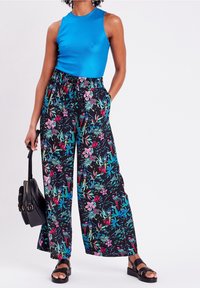 Blue sleeveless top, black floral wide-leg pants with pink and teal patterns. Model holds a black handbag, wearing black sandals.
