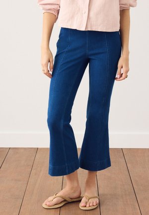 Frau trägt cropped ausgestellte blaue Jeans, ein hellrosa Hemd mit Knöpfen und beige Flip-Flops, steht auf einem Holzboden.