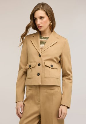 Kvinde iført tanfarvet cropped blazer med sorte knapper og matchende højtaljede bukser, der kigger en smule til siden mod en neutral baggrund.