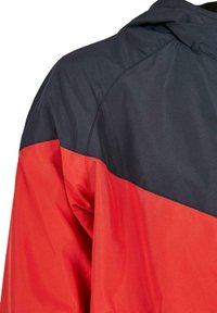 Több színű dzseki, amelynek fekete felső része és élénk piros alsó része van, sima, vízálló anyagból készült.