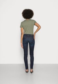 Vista trasera de una persona que lleva una camiseta de manga corta de color verde oliva y unos jeans skinny de color azul oscuro con costuras y bolsillos visibles.