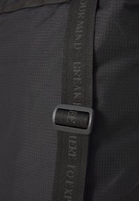 Black textured backpack material with a smooth strap featuring embossed lettering and a matte buckle detail.