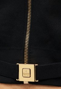 Μαύρο ριμπ ύφασμα με χρυστό μεταλλικό φερμουάρ και χρυσή μεταλλική λεπτομέρεια. Ο σχεδιασμός διαθέτει υφασμένο ζωνάρι για επιπλέον λεπτομέρεια.