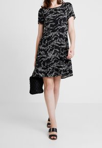 Svart klänning med vitt abstrakt mönster, korta ärmar, midjeindragning, flödande kjol, kombinerad med svart vävd väska och svarta klacksandaler.