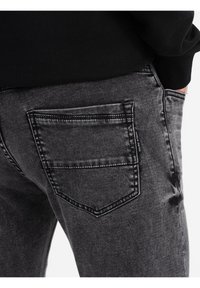 Jeans de mezclilla negra con un bolsillo trasero que tiene un contorno cosido, textura ligeramente desteñida y una sutil etiqueta de marca cerca de la costura del bolsillo.