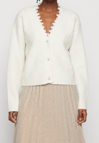 Biała kardigan z żeberkami, z diamentowym wzorem, z dekoltem w kształcie litery V, z zapięciem na guziki oraz koronkowym wykończeniem przy kołnierzu, zestawiona z plisowaną złotą spódnicą.