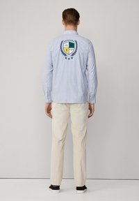 Blauw-wit gestreepte lange mouwen shirt met een schildlogo op de achterkant, gedragen met beige broeken en zwarte schoenen.