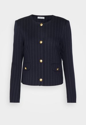 Vrouw draagt een navy pinstripe cropped blazer met gouden knopen, wit overhemd, blauwe cargobroek en zwarte puntige schoenen, staand tegen een grijze muur.