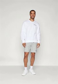 Sudadera blanca y pantalones cortos grises, ambos con acentos de logotipo. El modelo lleva zapatillas blancas y se encuentra frente a un fondo neutral.