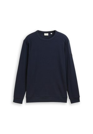 Tummansininen huppari, jossa on teksturoitu kangas, pyöreä kaula-aukko, raglan-hihat sekä resorit hihansuissa ja helmassa. Hänen hihaansa on lisätty pieni yksityiskohtapatch.