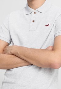 Šedé polo tričko s límcem, tříknoflíkovou légou a tmavě červeným logem. Krátké rukávy s jemnou texturou.