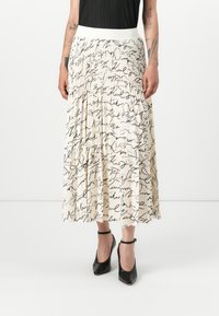 Jupe midi plissée avec une base crème ornée de motifs script écrits à la main en noir, taille élastique et tissu texturé. Chaussures à talons noirs.