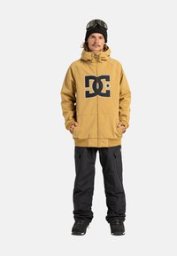 Geel ritsjack met zwart logo, gedragen met zwarte sneeuwbroeken en laarzen; heeft een capuchon en handschoenen, weergegeven op een neutrale achtergrond.