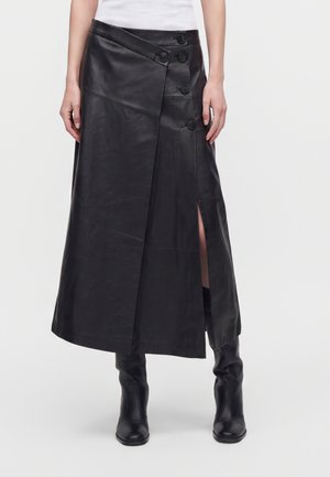 Femme portant une jupe midi portefeuille en cuir noir avec une fente latérale et des boutons, associée à des bottes noires montantes et un top blanc sans manches.