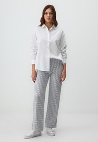Chemise blanche à manches longues avec des boutons, associée à un pantalon large gris à motifs. Le pantalon présente un design géométrique, complété par des baskets blanches.