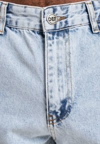 Lyseblå denimjeans med en falmet design, prydet med en sølvknapp med "DEF"-logo, kontrasterende sømmer og klassisk fem-lommers stil.