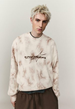 Junge Person mit kurzen blonden Haaren, die einen cremefarbenen Batikpullover mit grafischem Text und braune Hosen trägt, vor einem schlichten Hintergrund stehend.