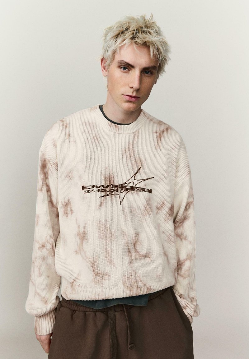 Jeune personne aux cheveux blonds courts portant un pull tie-dye crème avec un texte graphique et un pantalon marron, debout devant un fond uni.
