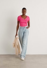 Roze v-hals t-shirt gecombineerd met lichtblauwe, wijde jeans. Het model houdt een crèmekleurige trui vast en draagt witte sneakers, met een overall gladde textuur.