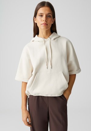 Jonge vrouw met lang bruin haar draagt een losse witte korte mouw hoodie en donkerbruine broek, staand tegen een effen lichte achtergrond.