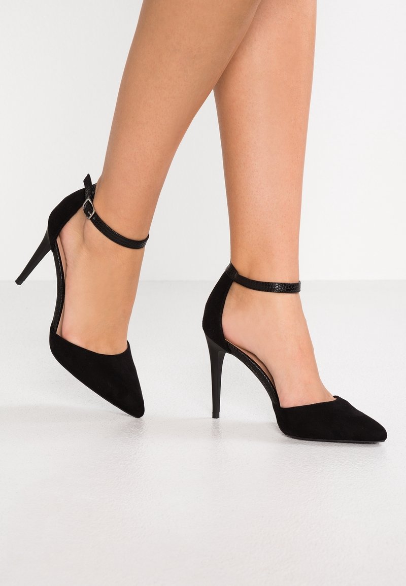 Escarpins en suede noir à bouts pointus, avec une bride autour de la cheville et un talon aiguille fin ; présentent des côtés ouverts et des accents texturés sur la bride.